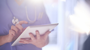 Female surgeon stood with her arms folded in hospital or surgery. She is wearing scrubs and has her stethoscope around her neck, the sun is bursting over her shoulder. She is looking at her patients records on her digital tablet and sending emails.